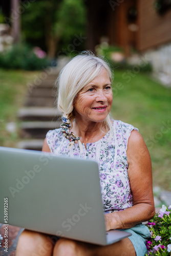 Senior woman using laptop and handling orders of her homegrown organic flowers and vegetables in garden, small business concept.
