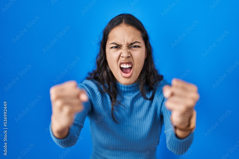 Young brazilian woman standing over blue isolated background angry and mad raising fists frustrated and furious while shouting with anger. rage and aggressive concept. Young brazilian woman standing over blue isolated background angry and mad raising fists frustrated and furious while shouting with anger. rage and aggressive concept.