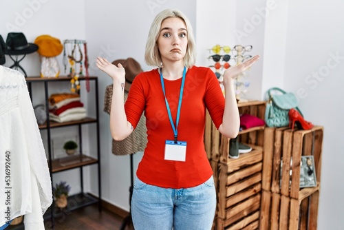 Young caucasian woman working as manager at retail boutique clueless and confused expression with arms and hands raised. doubt concept.