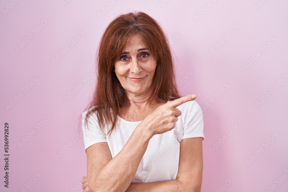 Middle age woman standing over pink background pointing with hand finger to the side showing advertisement, serious and calm face Middle age woman standing over pink background pointing with hand finger to the side showing advertisement, serious and calm face