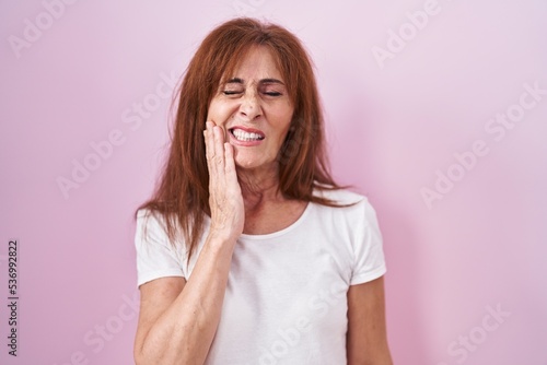 Middle age woman standing over pink background touching mouth with hand with painful expression because of toothache or dental illness on teeth. dentist