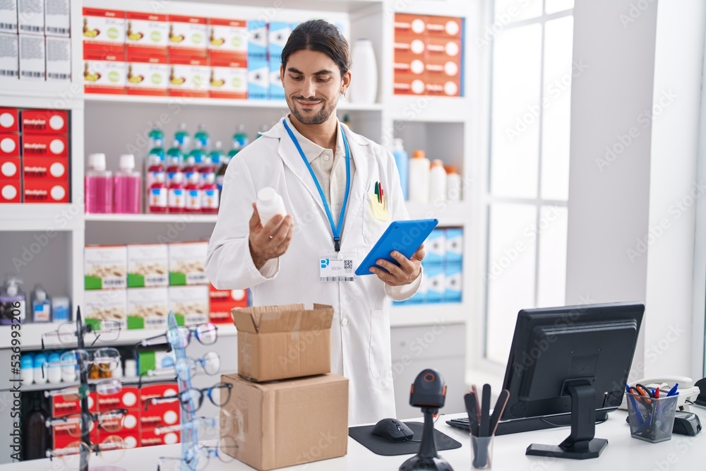 Young hispanic man pharmacist using touchpad holding pills bottle at pharmacy Young hispanic man pharmacist using touchpad holding pills bottle at pharmacy