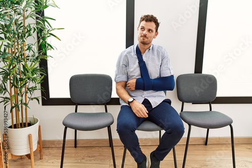 Handsome young man sitting at doctor waiting room with arm injury smiling looking to the side and staring away thinking.