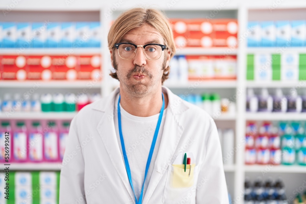 Caucasian man with mustache working at pharmacy drugstore puffing cheeks with funny face. mouth inflated with air, crazy expression.