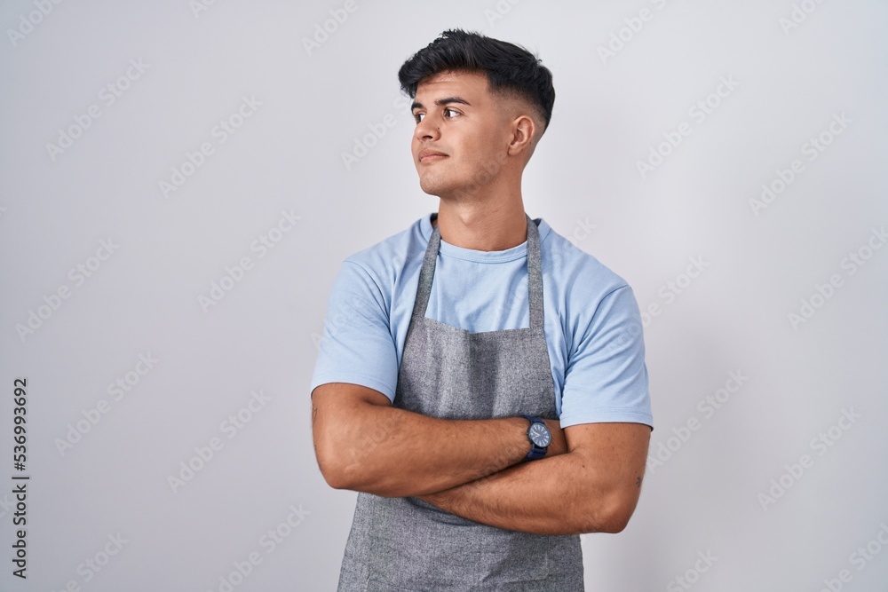 Hispanic young man wearing apron over white background looking to the side with arms crossed convinced and confident Hispanic young man wearing apron over white background looking to the side with arms crossed convinced and confident