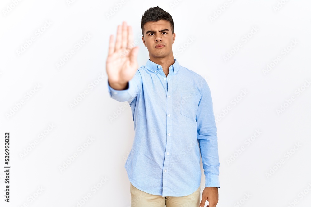 Young hispanic man wearing business shirt standing over isolated background doing stop sing with palm of the hand. warning expression with negative and serious gesture on the face. Young hispanic man wearing business shirt standing over isolated background doing stop sing with palm of the hand. warning expression with negative and serious gesture on the face.