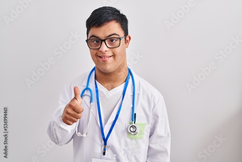 Young hispanic man with down syndrome wearing doctor uniform and stethoscope smiling happy and positive  thumb up doing excellent and approval sign