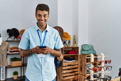 Young hispanic man working as shop assistant using smartphone at retail shop