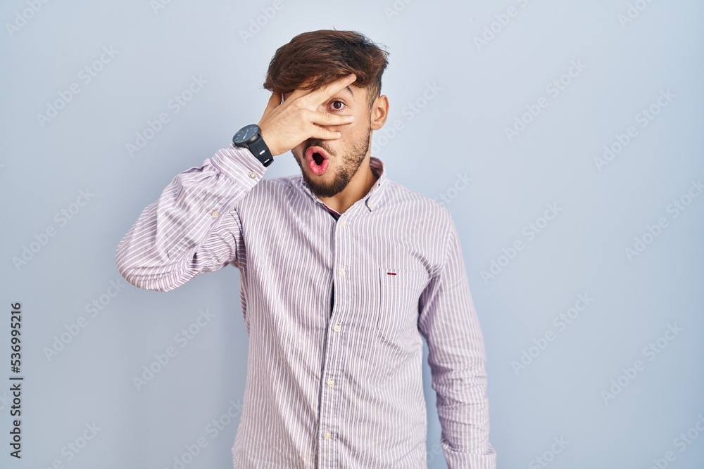 Arab man with beard standing over blue background peeking in shock covering face and eyes with hand, looking through fingers with embarrassed expression. Arab man with beard standing over blue background peeking in shock covering face and eyes with hand, looking through fingers with embarrassed expression.