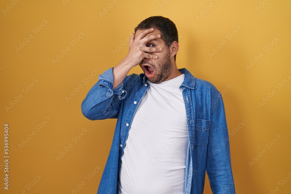 Hispanic man standing over yellow background peeking in shock covering face and eyes with hand, looking through fingers with embarrassed expression. Hispanic man standing over yellow background peeking in shock covering face and eyes with hand, looking through fingers with embarrassed expression.