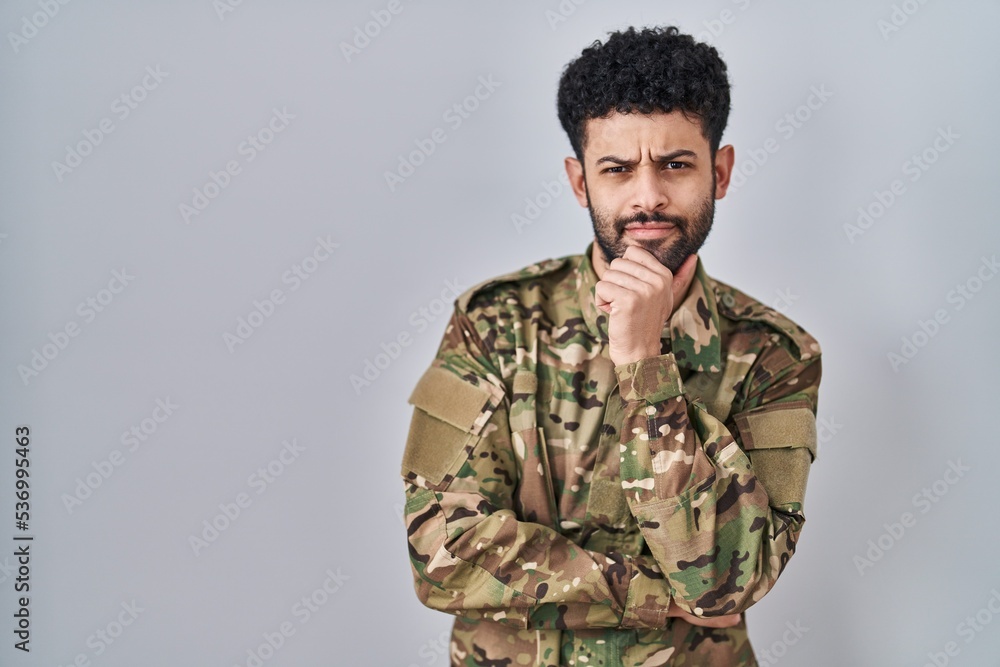 Arab man wearing camouflage army uniform looking confident at the camera smiling with crossed arms and hand raised on chin. thinking positive.