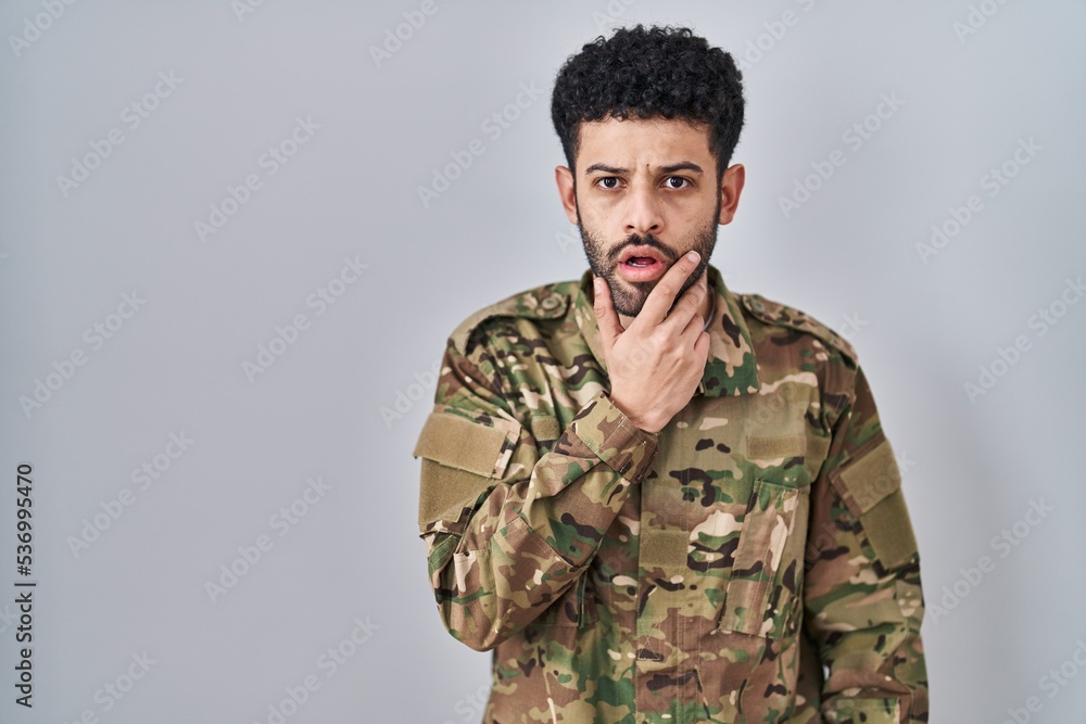 Arab man wearing camouflage army uniform looking fascinated with disbelief, surprise and amazed expression with hands on chin