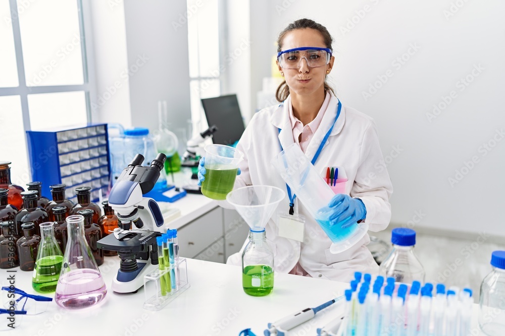Fototapeta premium Young hispanic woman working at scientist laboratory puffing cheeks with funny face. mouth inflated with air, catching air.