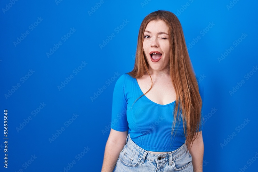 Redhead woman standing over blue background winking looking at the camera with sexy expression, cheerful and happy face. Redhead woman standing over blue background winking looking at the camera with sexy expression, cheerful and happy face.