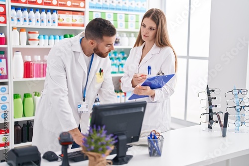 Man and woman pharmacists using computer writing on document at pharmacy