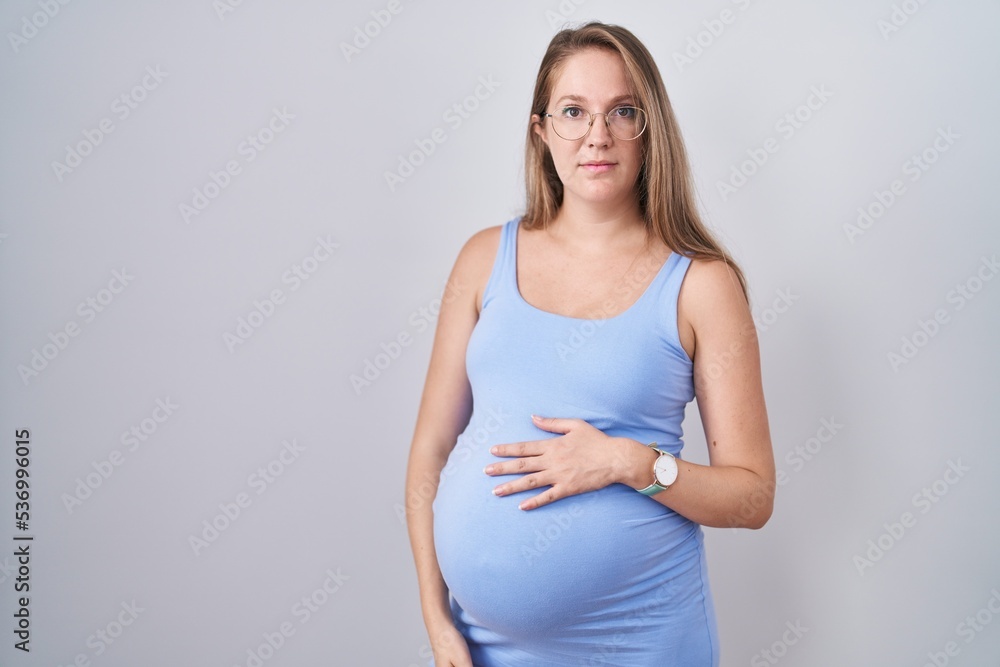 Young pregnant woman standing over white background relaxed with serious expression on face. simple and natural looking at the camera. Young pregnant woman standing over white background relaxed with serious expression on face. simple and natural looking at the camera.
