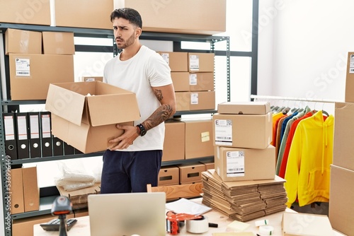 Young hispanic man working at small business ecommerce clueless and confused expression. doubt concept.