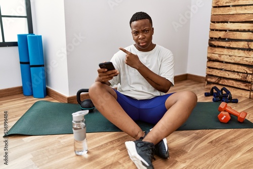 Young african man sitting on training mat at the gym using smartphone pointing aside worried and nervous with forefinger  concerned and surprised expression