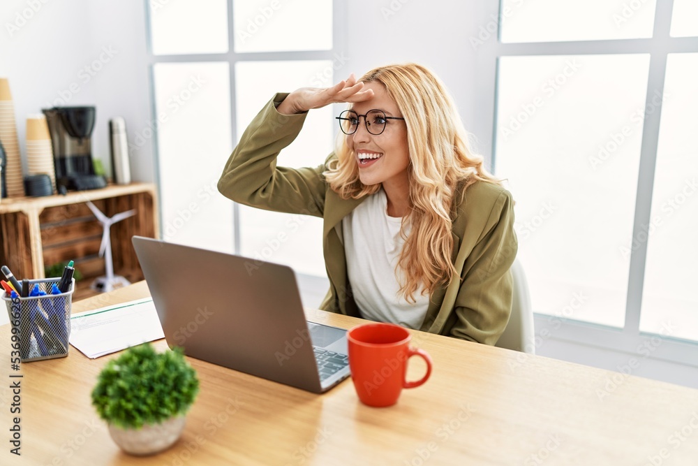 Beautiful blonde woman working at the office with laptop very happy and smiling looking far away with hand over head. searching concept. Beautiful blonde woman working at the office with laptop very happy and smiling looking far away with hand over head. searching concept.