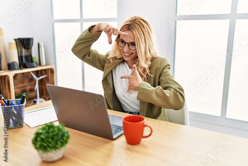 Beautiful blonde woman working at the office with laptop smiling making frame with hands and fingers with happy face. creativity and photography concept.