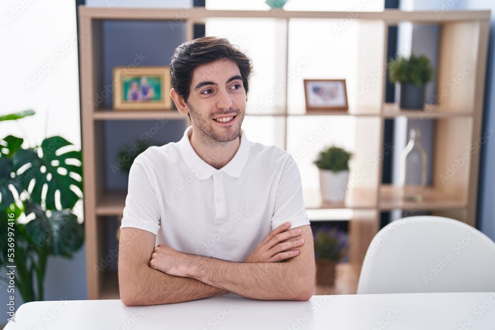 Young hispanic man smiling confident sitting on table with arms crossed gesture at home Young hispanic man smiling confident sitting on table with arms crossed gesture at home