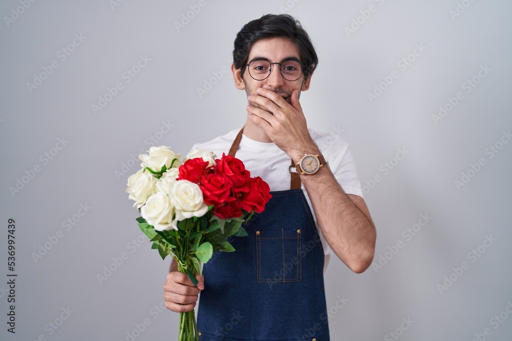 Young hispanic man holding bouquet of white and red roses laughing and embarrassed giggle covering mouth with hands, gossip and scandal concept Young hispanic man holding bouquet of white and red roses laughing and embarrassed giggle covering mouth with hands, gossip and scandal concept