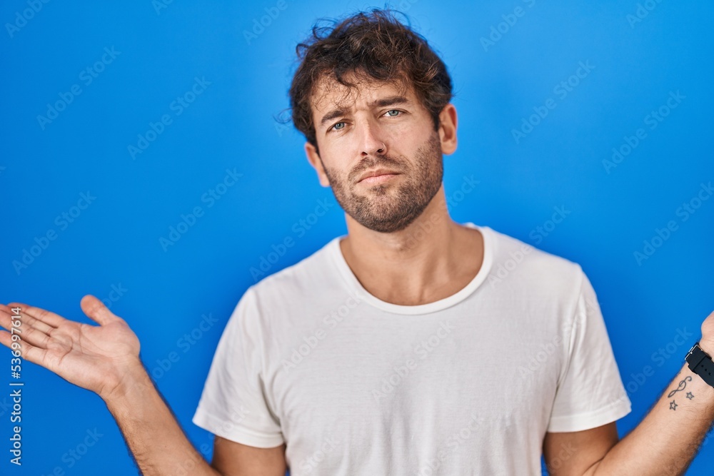 Fototapeta premium Hispanic young man standing over blue background clueless and confused expression with arms and hands raised. doubt concept.