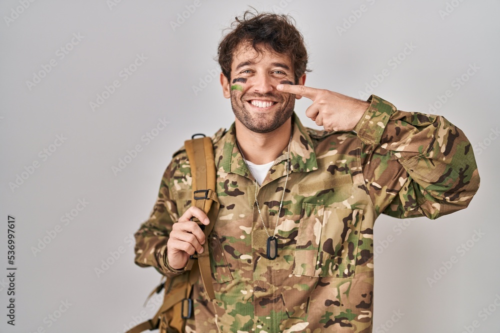 Hispanic young man wearing camouflage army uniform pointing with hand finger to face and nose, smiling cheerful. beauty concept Hispanic young man wearing camouflage army uniform pointing with hand finger to face and nose, smiling cheerful. beauty concept
