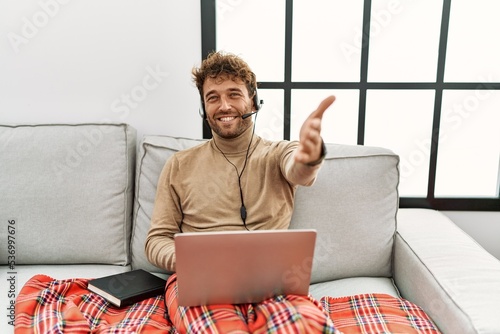 Young handsome man with beard wearing operator headset working from home smiling friendly offering handshake as greeting and welcoming. successful business.