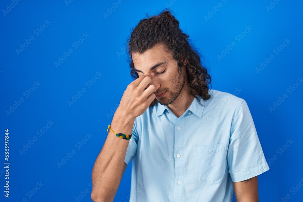 Young hispanic man standing over blue background tired rubbing nose and eyes feeling fatigue and headache. stress and frustration concept. Young hispanic man standing over blue background tired rubbing nose and eyes feeling fatigue and headache. stress and frustration concept.