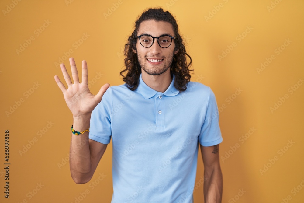 Young hispanic man standing over yellow background showing and pointing up with fingers number five while smiling confident and happy. Young hispanic man standing over yellow background showing and pointing up with fingers number five while smiling confident and happy.