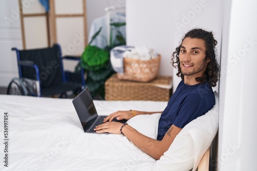 Young hispanic man disabled using laptop sitting on bed at bedroom