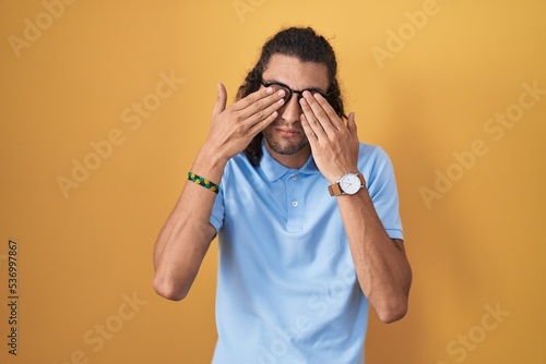 Young hispanic man standing over yellow background rubbing eyes for fatigue and headache, sleepy and tired expression. vision problem