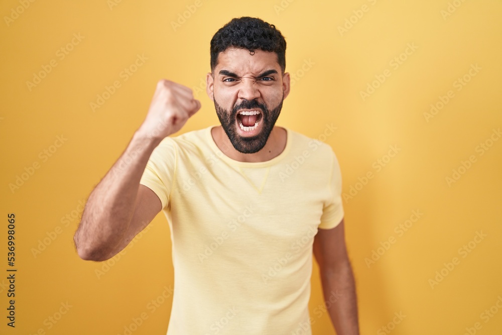 Hispanic man with beard standing over yellow background angry and mad raising fist frustrated and furious while shouting with anger. rage and aggressive concept. Hispanic man with beard standing over yellow background angry and mad raising fist frustrated and furious while shouting with anger. rage and aggressive concept.
