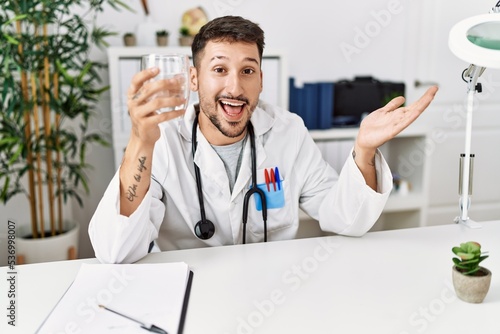 Young doctor holding glass of water celebrating achievement with happy smile and winner expression with raised hand