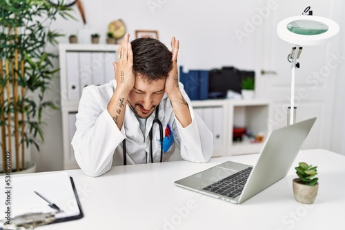 Young doctor working at the clinic using computer laptop suffering from headache desperate and stressed because pain and migraine. hands on head.