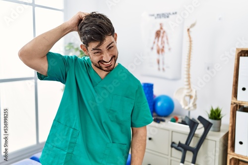 Young man with beard working at pain recovery clinic confuse and wonder about question. uncertain with doubt  thinking with hand on head. pensive concept.