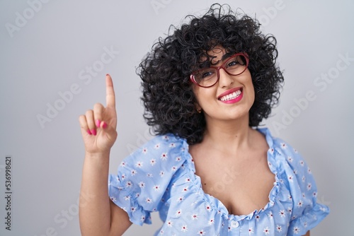 Young brunette woman with curly hair wearing glasses over isolated background pointing finger up with successful idea. exited and happy. number one.