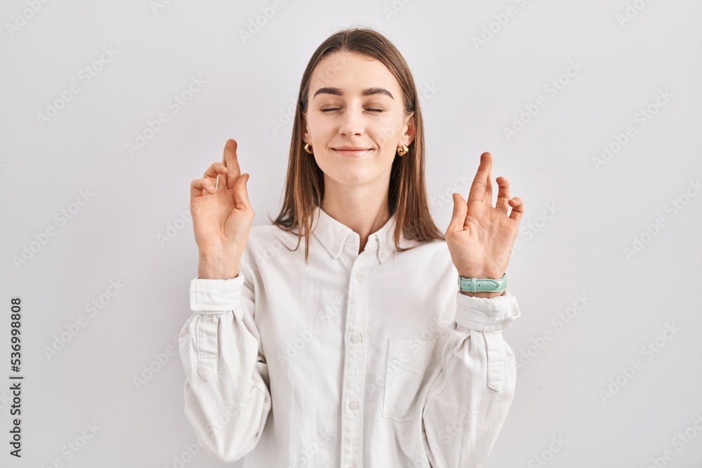 Young caucasian woman standing over isolated background gesturing finger crossed smiling with hope and eyes closed. luck and superstitious concept. Young caucasian woman standing over isolated background gesturing finger crossed smiling with hope and eyes closed. luck and superstitious concept.