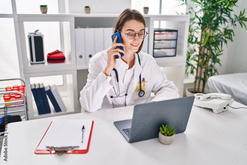 Young caucasian woman doctor using laptop talking on smartphone at clinic Young caucasian woman doctor using laptop talking on smartphone at clinic