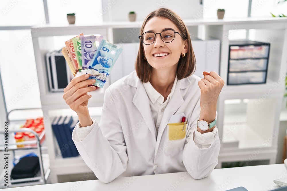 Young caucasian doctor woman holding canadian dollars banknotes screaming proud, celebrating victory and success very excited with raised arms Young caucasian doctor woman holding canadian dollars banknotes screaming proud, celebrating victory and success very excited with raised arms