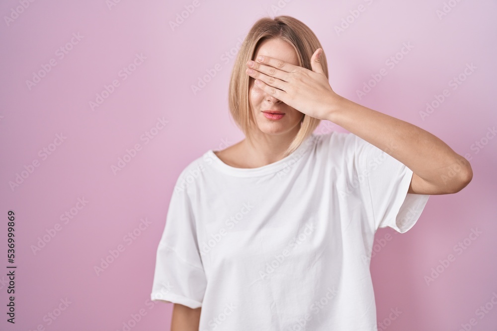 Young caucasian woman standing over pink background covering eyes with hand, looking serious and sad. sightless, hiding and rejection concept Young caucasian woman standing over pink background covering eyes with hand, looking serious and sad. sightless, hiding and rejection concept