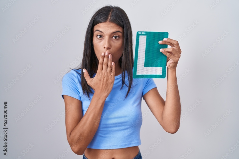 Brunette young woman holding l sign for new driver covering mouth with hand, shocked and afraid for mistake. surprised expression Brunette young woman holding l sign for new driver covering mouth with hand, shocked and afraid for mistake. surprised expression