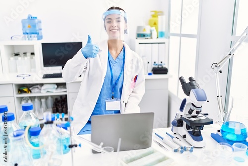 Young hispanic woman wearing scientist uniform smiling happy and positive, thumb up doing excellent and approval sign