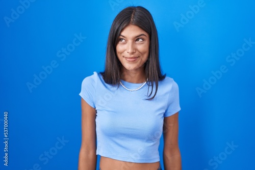 Brunette young woman standing over blue background smiling looking to the side and staring away thinking.
