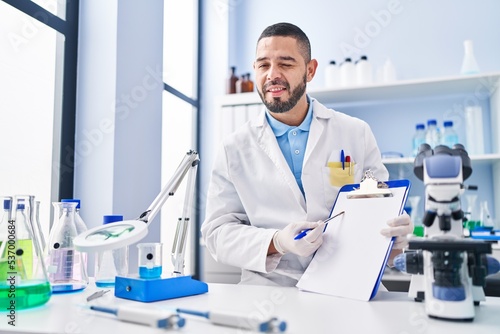 Hispanic man working at scientist laboratory holding blank clipboard winking looking at the camera with sexy expression  cheerful and happy face.