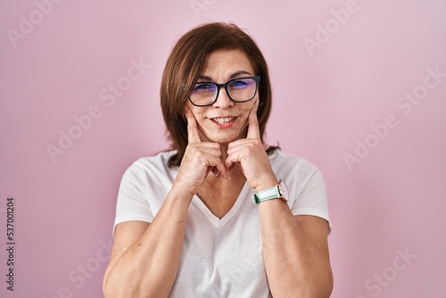 Middle age hispanic woman standing over pink background smiling with open mouth, fingers pointing and forcing cheerful smile