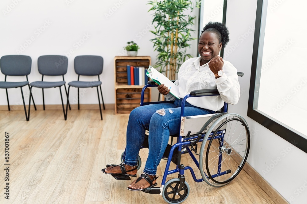 Young black woman sitting on wheelchair at waiting room very happy and excited doing winner gesture with arms raised, smiling and screaming for success. celebration concept. Young black woman sitting on wheelchair at waiting room very happy and excited doing winner gesture with arms raised, smiling and screaming for success. celebration concept.