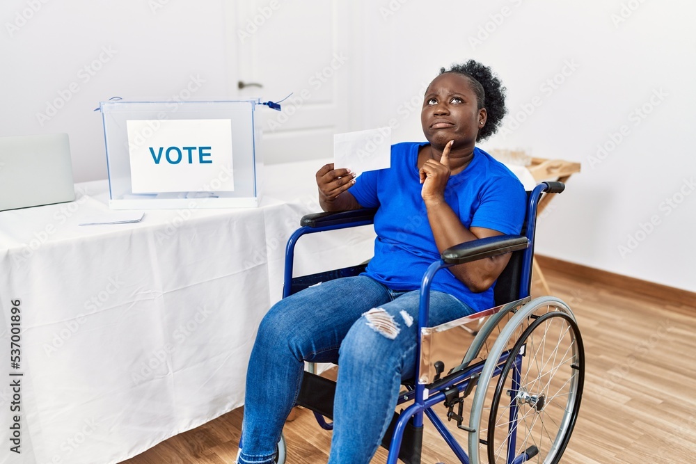 Young african woman sitting on wheelchair voting putting envelop in ballot box thinking concentrated about doubt with finger on chin and looking up wondering Young african woman sitting on wheelchair voting putting envelop in ballot box thinking concentrated about doubt with finger on chin and looking up wondering