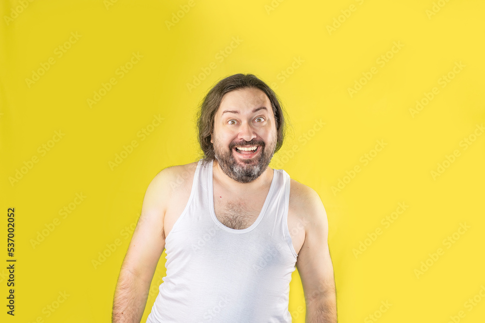 Obraz premium A joyful, unshaven man in a white undershirt stands and smiles exaggeratedly. Unemployed slovenly dressed man from Eastern Europe.